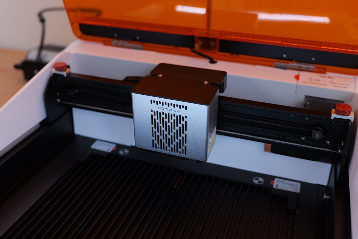 Buồng khắc kín với đầu laser trên ray và bàn tổ ong — có nút dừng khẩn hai bên.